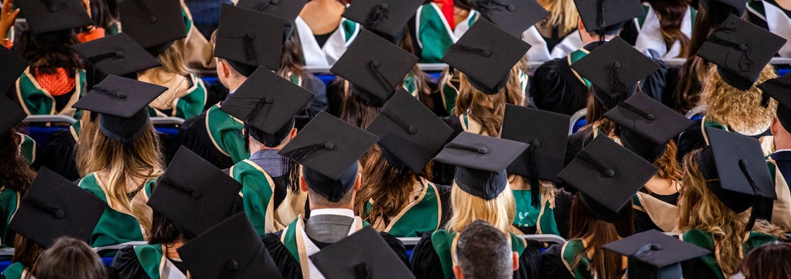 A shot of students in the Aula, sitting facing the stage.  The shot shows the back of their heads, master's robes and mortar boards