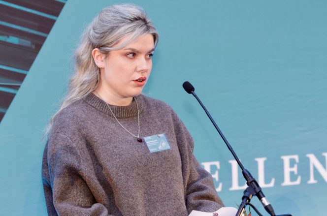 student Edith May Hand, pictured speaking at a microphone wearing a grey jumper against a blue background