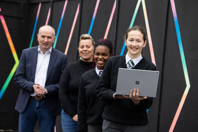 4 poeple looking at the camera smiling. 1 man in a suit, 1 woman, 2 girls in school uniforms one holding a laptop