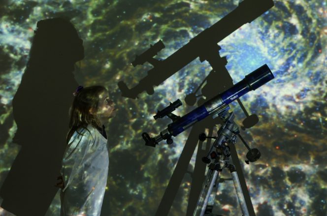 small girl with a telescope surrounded by a projection of space and stars