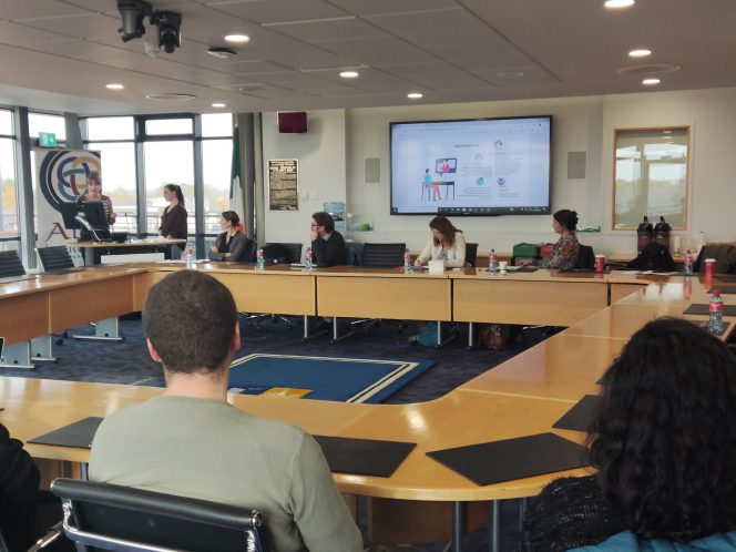 People sitting around a boardroom Holly and Neasa behind the podium presenting with a screen in the background showing information on digital wealth