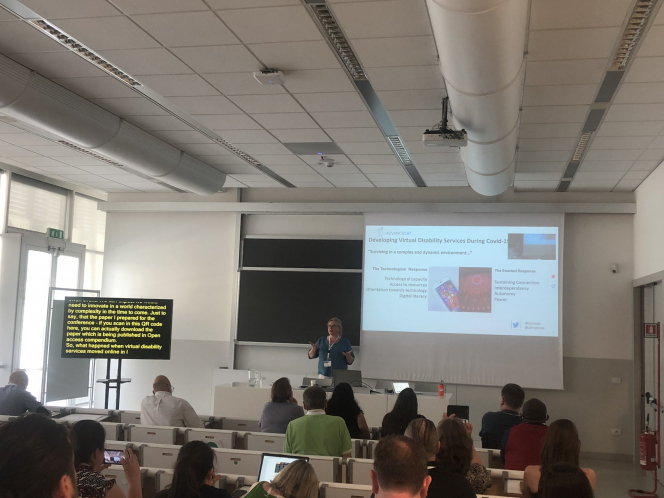 Joan O Donnell standing in front of a room presenting with English subtitles on screen to the left of the image with a screen behind her reading: ADVANCE CRT. Developing virtual disability during Covid-19