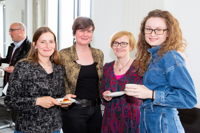 4 women smiling at the camera woman on the far left holding a plate of food woman on the far right holding a mug