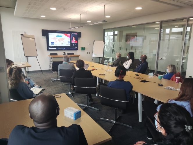 Image from the back of the room showing people looking towards the screen. The screen shows Natasha Layton presenting her slides.