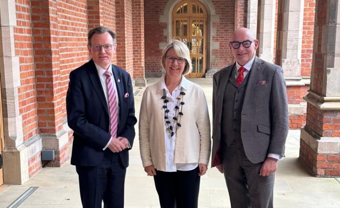 1 women in the middle in a white jacket, and black pants, with 2 other men. Man to the left in dark suit, white shirt and pink tie, man to the right, bald and glasses with a grey suit with red tie