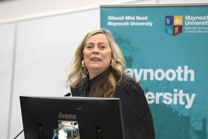 Woman with blond hair standing behind a lectern and in front of a popup of Maynooth University
