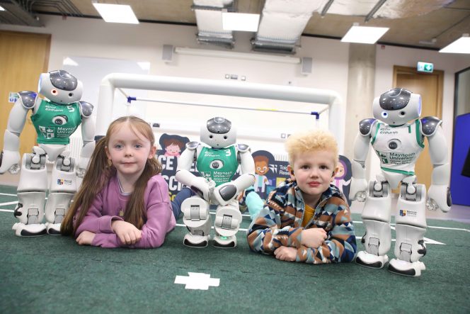 Little girl in pink and little boy with blond curly hair pose with three soccer robots