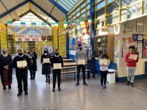 a bright interior image of the school with lots of windows and glass paneled roof, six students are holding certificates and three staff members stand behind them. There is bunting in the background with Irish flags displayed. 