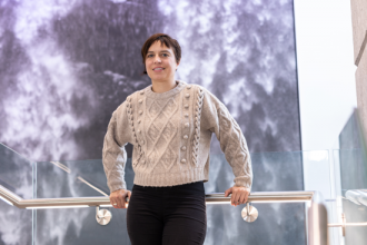 Elisa Fadda faces the camera, standing on a stair landing,  in front of a wall hanging showing flowing water