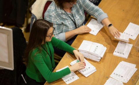 2 women - women on the left with a green top, sitting at a desk counting ballot papers, woman on the right wearing a blue/white blouse, also counting ballot papers.