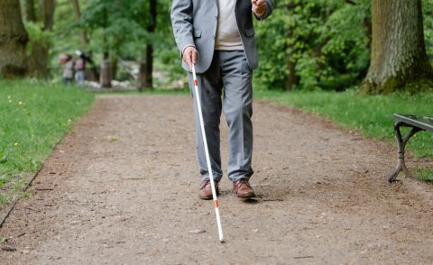 Man in a grey suit with a white cane walking through a park