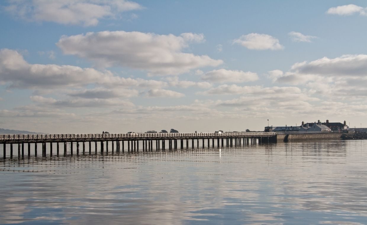 A day time shot of the wooden bridge linking Clontarf to Bull Island, the sea below has barely any waves and is reflecting the clouds and blue sky like a mirror. 