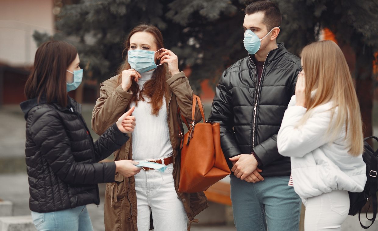 3 women and 1 man talking to each other wearing covid masks. Black jackets. blue jeans, white jacket and brown jacket 