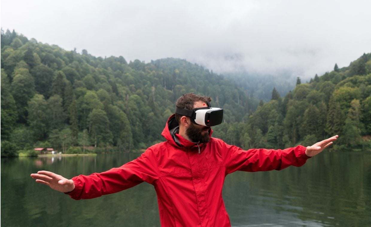 Man with a beard wearing a red jacket and VR goggles standing in front of a lake with forestry on both sides