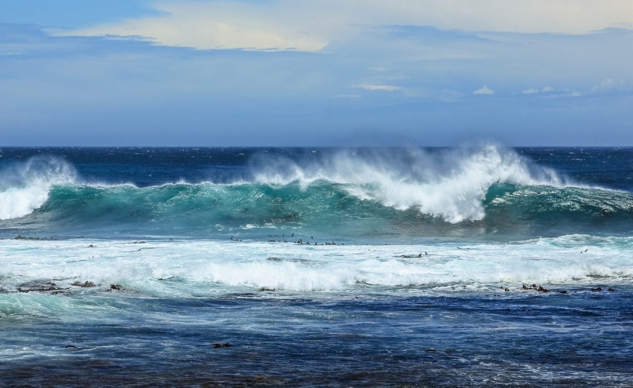 Waves breaking with foam under a blue sky with calm blue water closer to shore