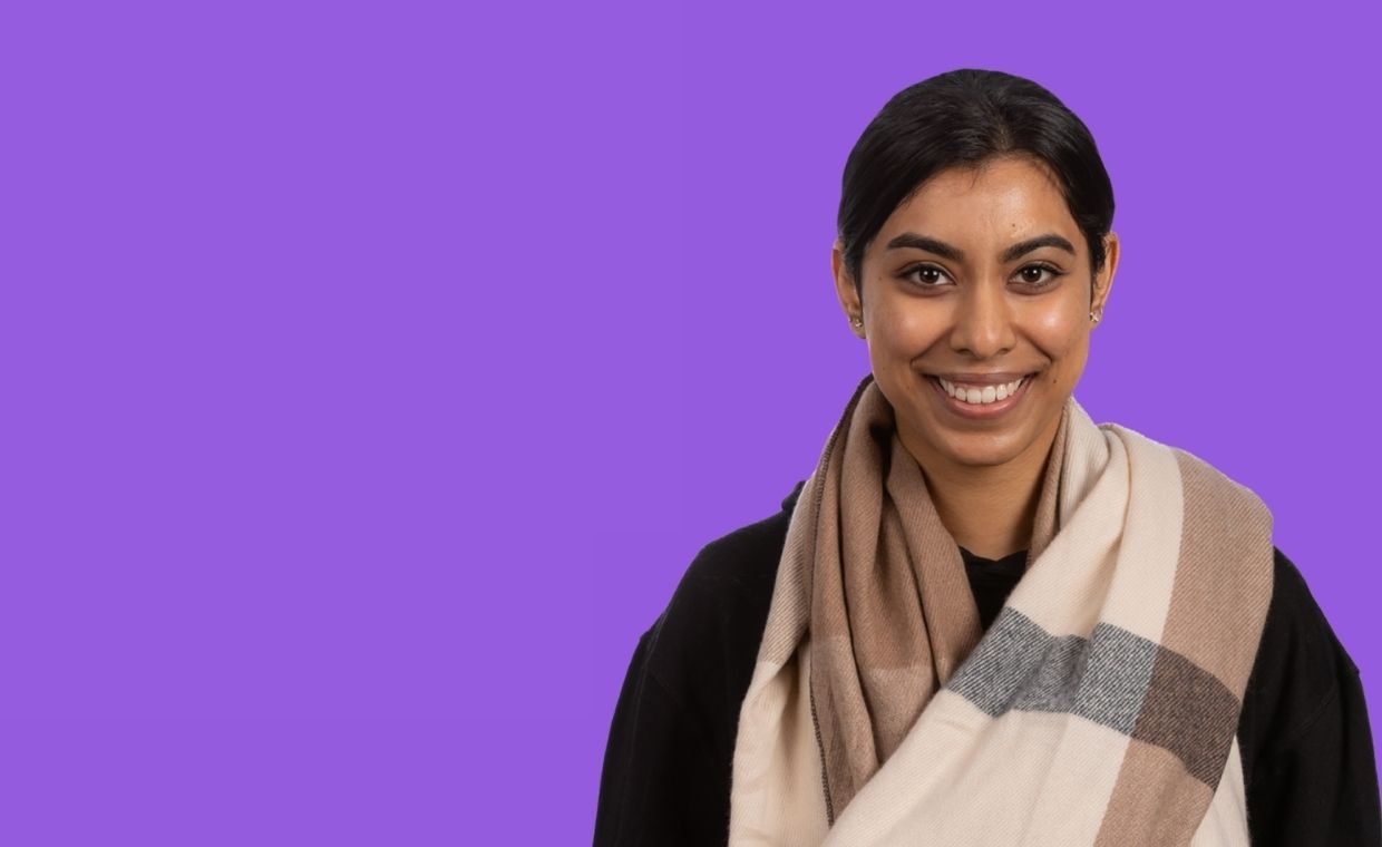 A postgraduate student wearing a beige tartan scarf smiles.  The background is a solid block of bright purple