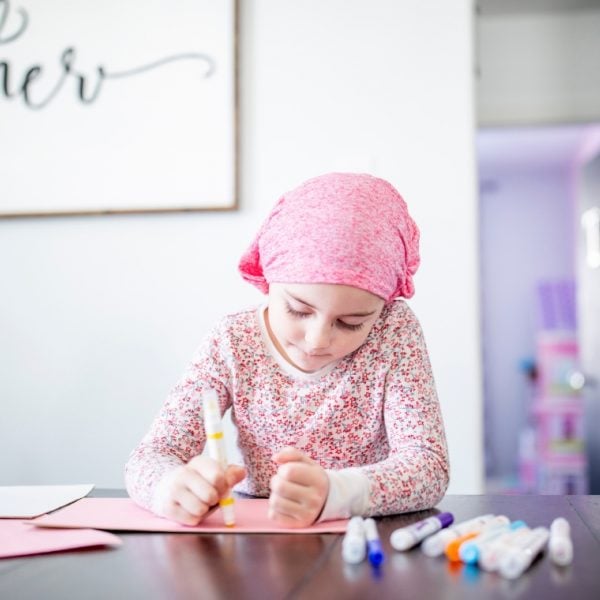 Little girl wearing a pink headscarf sitting at a table drawing with magic markers