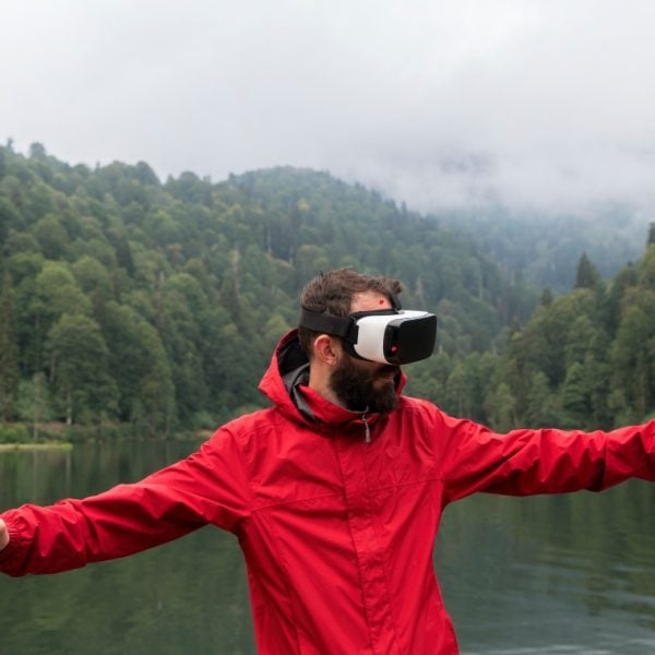 Man with a beard wearing a red jacket and VR goggles standing in front of a lake with forestry on both sides