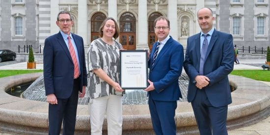 Group of three men and one woman standing in front of a fountain, holding a large certificate