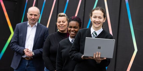 4 poeple looking at the camera smiling. 1 man in a suit, 1 woman, 2 girls in school uniforms one holding a laptop