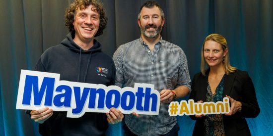 Man on left in navy hoodie, Man in middle in check pattern shirt and woman on the right wearing black top. All holding sign 'Maynooth #Alumni