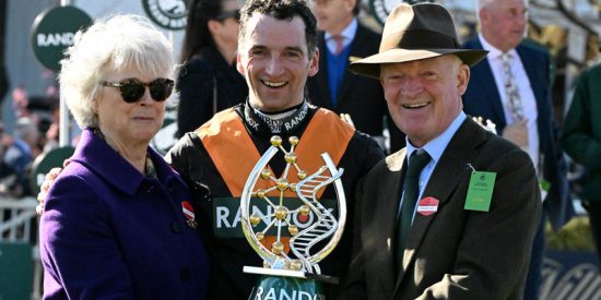 A medium shot shows three smiling people holding a trophy. In the center is Patrick Mullins, wearing racing silks of black with orange accents To his left is his mother, Jackie Mullins. She wears a purple coat and a gold bracelet. To the right of Patrick is trainer and Patrick’s father, Willie Mullins, wearing a brown fedora, a brown tweed jacket, a light blue collared shirt, and a dark green tie. He is also smiling at the camera