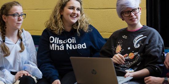 3 students sitting around a laptop.  One wears a sweater with 'OllScoil Mhá Nuad'