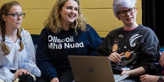 3 students sitting around a laptop. One wears a sweater with 'OllScoil Mhá Nuad'