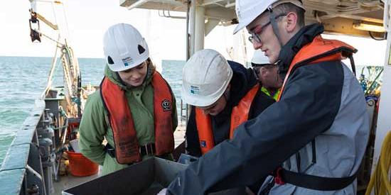 Three research students in white hard hats and life jackets inspecting samples on a boat at sea