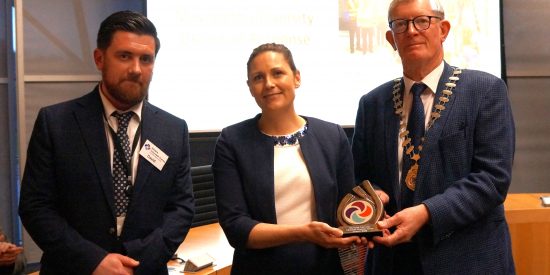 Three people at an award ceremony with two men on either side of woman holding an award.