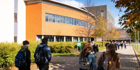 Four students, two men and two women, walking along a pathway beside an orange University building