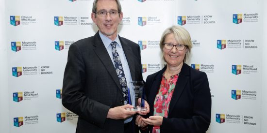 Tall man in a dark suit receiving an award from a woman in a black jacket and red blouse