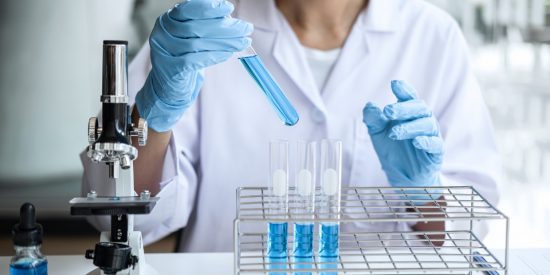Scientist in a white lab coat and blue gloves, holding a test tube with blue liquid beside a microscope