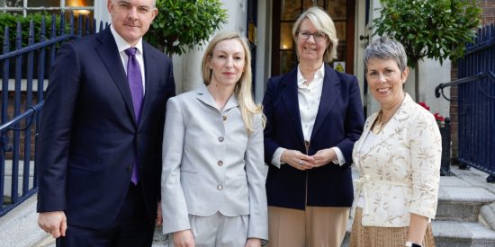 Man in blue suit, woman in light grey suit, woman in navy jacket and woman in white jacket standing in front of a doorway