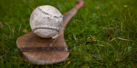Picture of a white sliotar sitting on top of a weathered hurl on grass