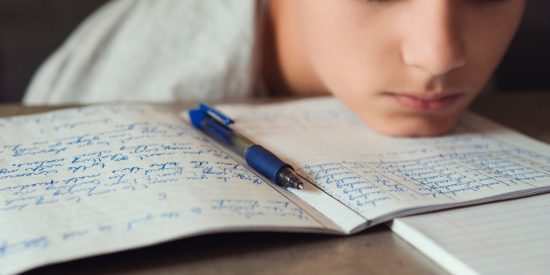 Child with sad face and head resting on table. Book with writing and a pen