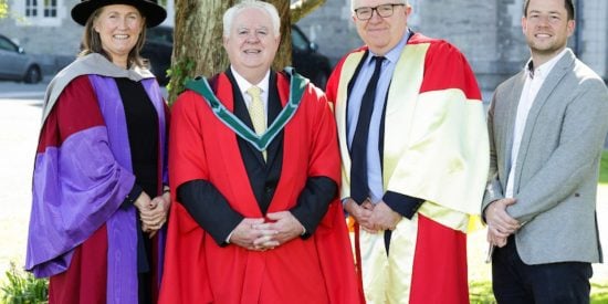 Woman and two men in academic robes and a man in a grey jacket stand in front of a tree