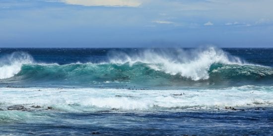 Waves breaking with foam under a blue sky with calm blue water closer to shore