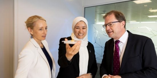 Woman in white jacket, woman in black jacket and white head cover holding a petri dish watched by a man in a black jacket