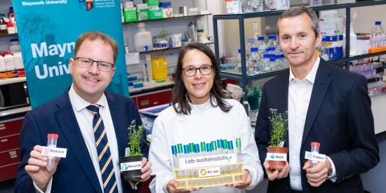 Two men and a woman in a white coat holding a rack of test tubes in a laboratory