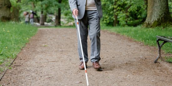 Man in a grey suit with a white cane walking through a park