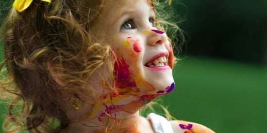 Photo of female child, coverd in paint with a yellow ribbon in her hair, looking very happy