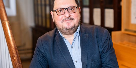 man with glasses and beard wearing a navy suit jacket and baby blue shirt posing against wooden stair rail