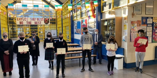 a bright interior image of the school with lots of windows and glass paneled roof, six students are holding certificates and three staff members stand behind them. There is bunting in the background with Irish flags displayed. 