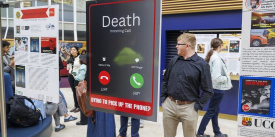 The image shows a poster with the words "Death Incoming Call" in large text. Below, there are two phone icons labeled "LIFE" and "RIP," and the question "DYING TO PICK UP THE PHONE?" The is part of the Road Safety Reimagined exhibition, with other posters and people visible in the background. 