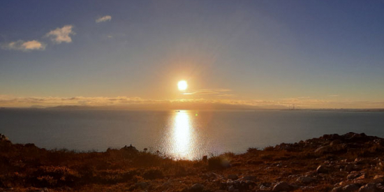 A view out a very calm sea from the land, the sun is setting and its light is stretching in a path across the water