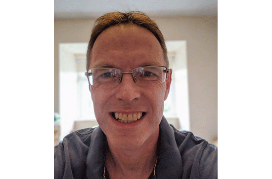A portrait of Donal O'Neil, smiling to the camera, Donal is wearing a grey collared shirt. 