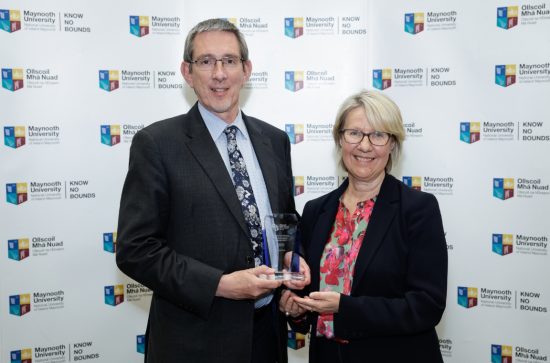 Tall man in a dark suit receiving an award from a woman in a black jacket and red blouse