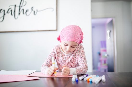 Little girl wearing a pink headscarf sitting at a table drawing with magic markers