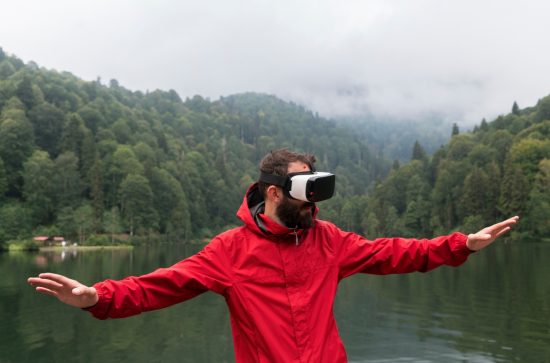 Man with a beard wearing a red jacket and VR goggles standing in front of a lake with forestry on both sides